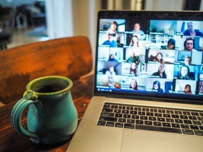 A mug next to laptop displaying a group of people on a virtual video call on a wooden table and wooden chair