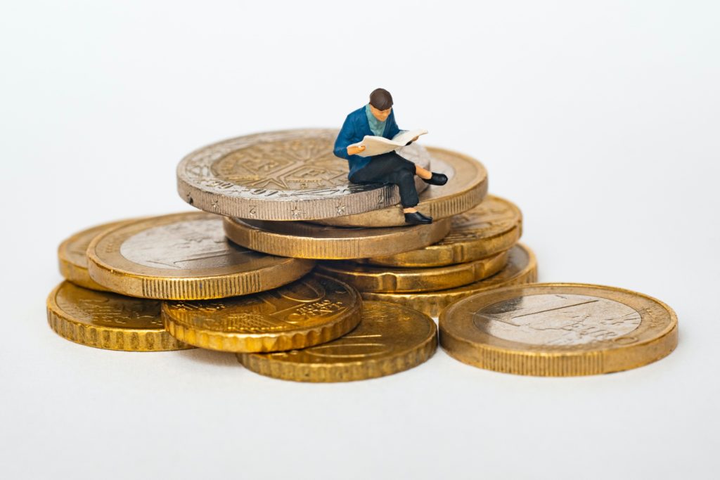 A close up view of a tiny toy of a man reading a book while sitting on a stack of gold euro coins