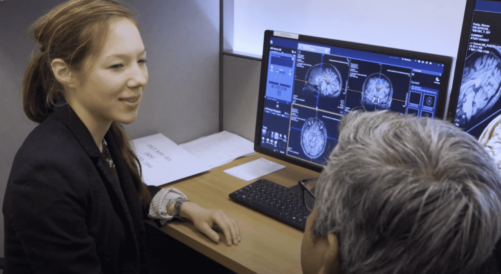 A woman speaking to a surgeon at a computer desk with brain X-Rays displaying on the monitors