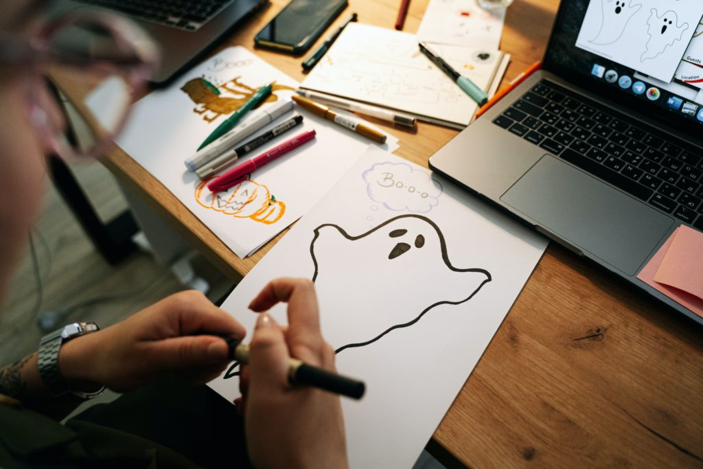 A woman drawing a flying ghost with a black marker while using her laptop as a reference alongside other drawings such as pumpkins and words such as boo