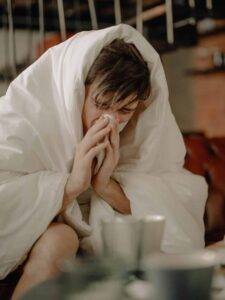 A man blowing his nose while covered in a white blanket sitting in a bed