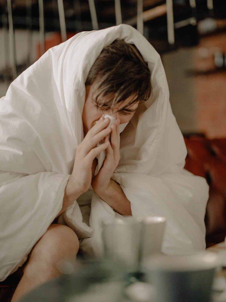A man blowing his nose while covered in a white blanket sitting in a bed
