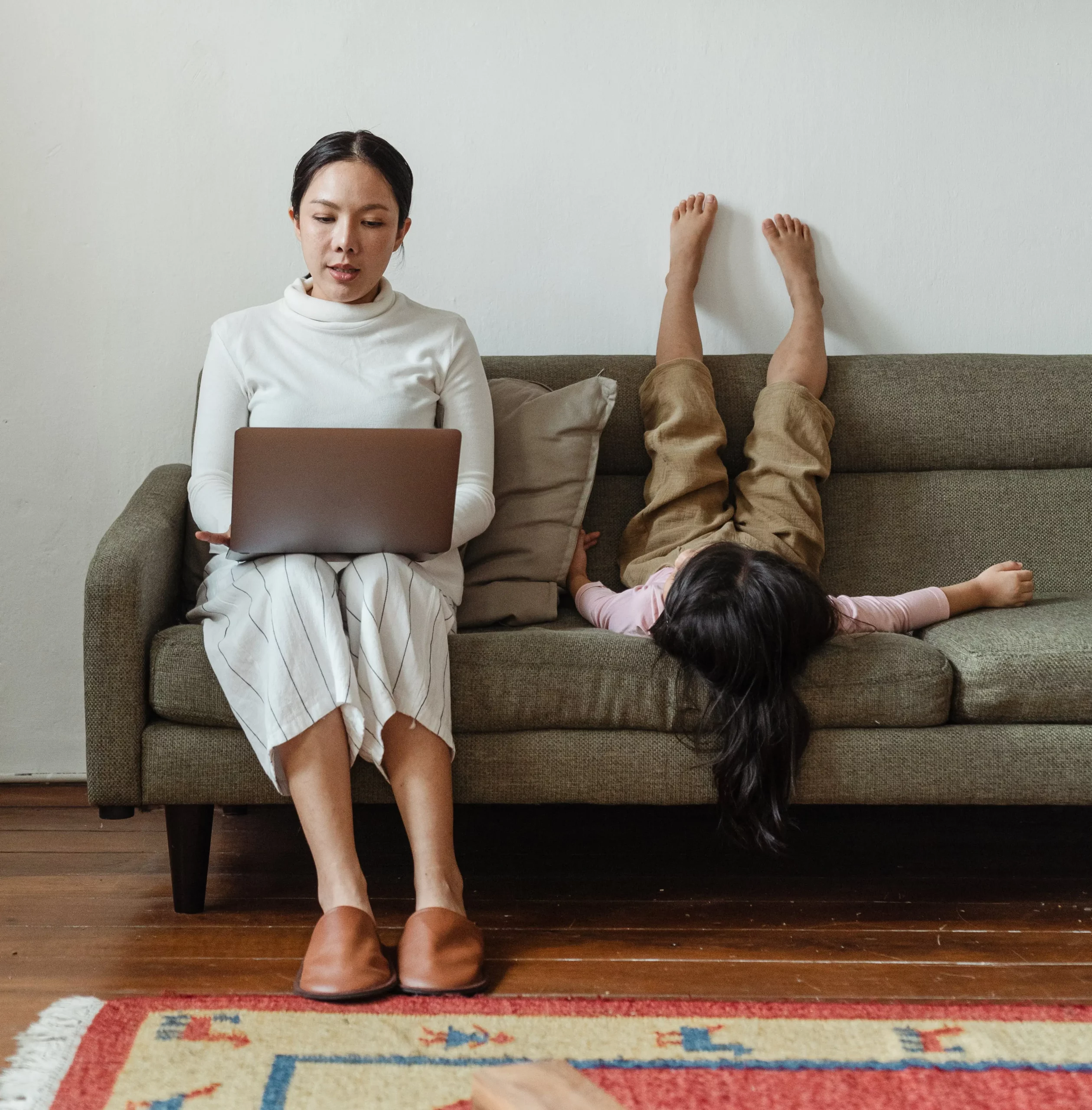 A woman doing remote working on a laptop next to a child on a couch.