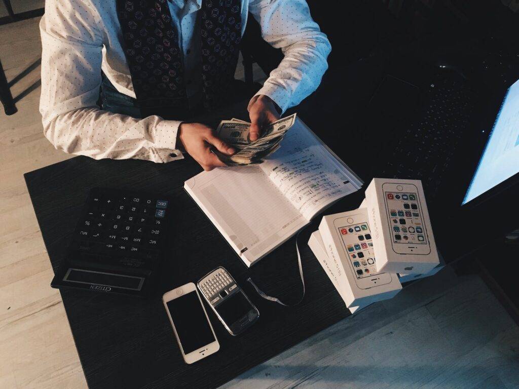 a person counting money at a desk, sitting by a calculator, phones, book, and laptop, representing the importance of Audit Documentation 