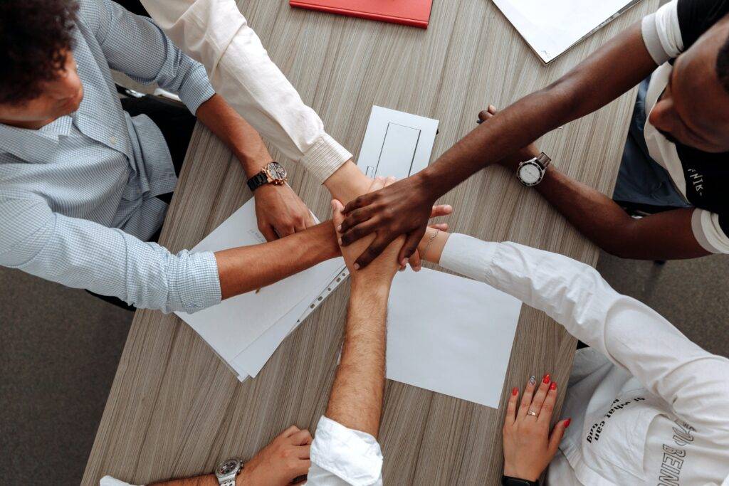 a number of hands joined together at the center of a table, representing the teamwork benefits of white paper writing