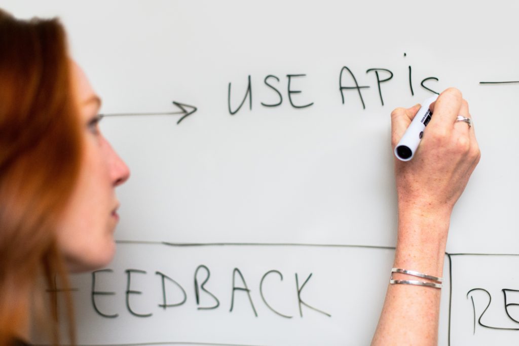 A woman writing the acronym "APIs" on a whiteboard using a black board marker